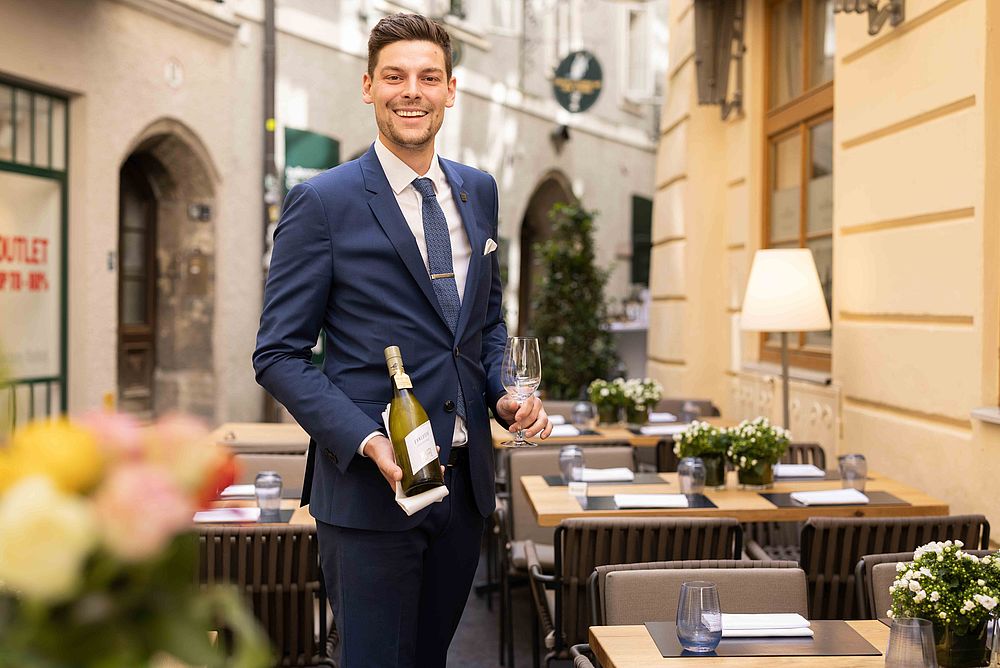 [Translate to Italienisch:] Ein Mann präsentiert einen edlen Champagner auf der Terrasse des Gasthof Goldgasse